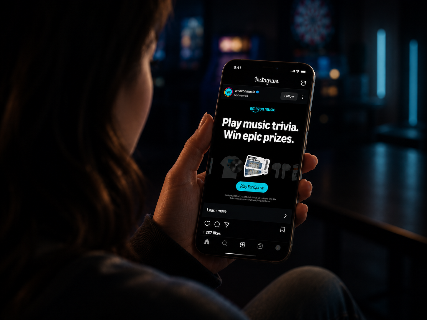 A person holding a phone in a dark room, viewing a sponsored Amazon Music Instagram post for FanQuest — 'Play music trivia. Win epic prizes.'